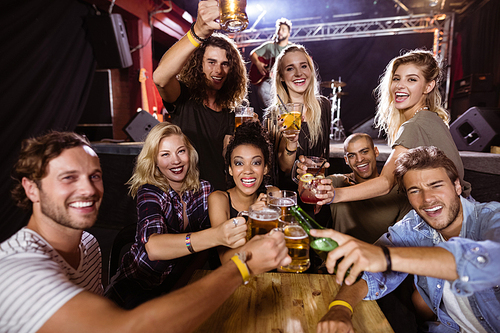 Portrait of cheerful friends toasting drink at table with performer singing on stage in nightclub