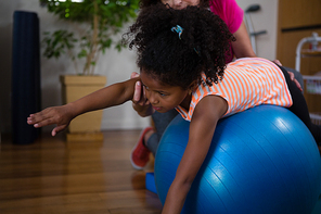 Female physiotherapist helping girl patient in performing exercise on fitness ball in clinic