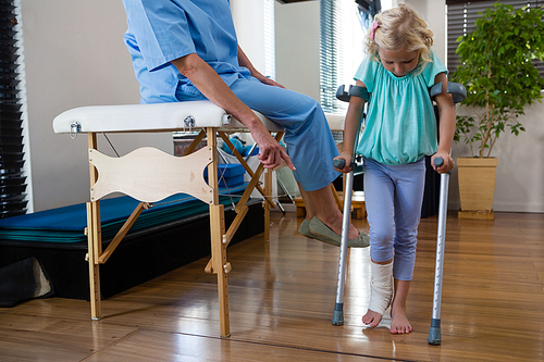 Physiotherapist assisting girl patient to walk with crutches in clinic