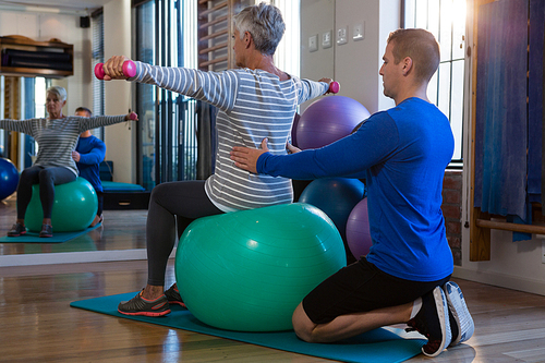Physiotherapist assisting senior woman on exercise ball and dumbbells at clinic