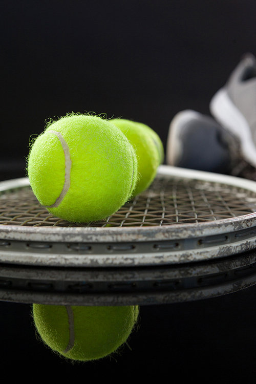 Close up of fluorescent yellow tennis balls on racket by sports shoes with reflection against black background