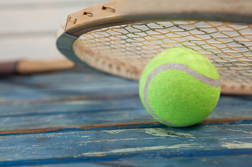 Close up of wooden tennis racket leaning on fluorescent yellow ball over blue table