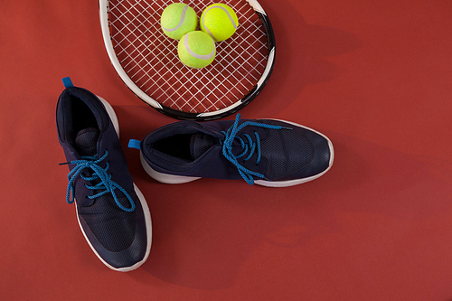 Overhead view of blue sports shoes by tennis racket and balls on maroon background