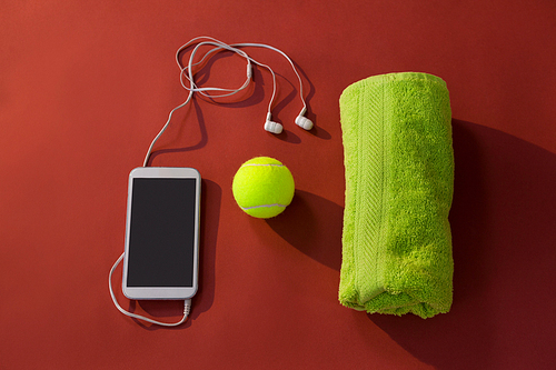 Overhead view of tennis ball amidst napkin and mobile phone with in-ear headphones on maroon background