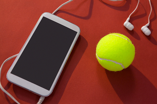 High angle view of tennis ball and mobile phone with in-ear headphones on maroon background