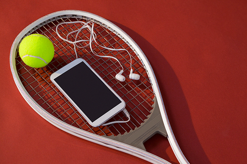 High angle view of mobile phone with in-ear headphones and ball on tennis racket over maroon background