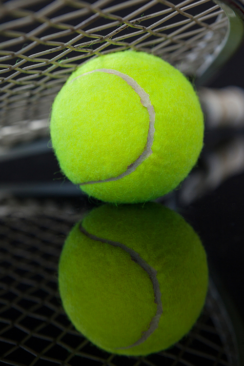 Close up of tennis racket on fluorescent yellow ball with reflection against black background