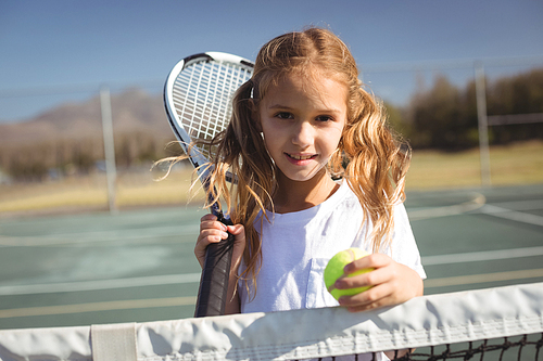 Portrait of girl holding tennis racket and ball while standing by net on court