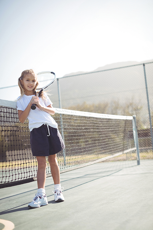 Full length portrait of girl holding tennis racket while sitting by net on sunny day