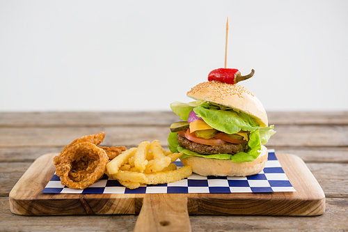 Close up of hamburger with onion rings and french fries on cutting board at table against wall