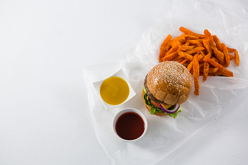 Overhead view of hamburger by French fries and dips in bowl on white table