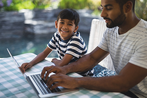 Portrait of smiling boy sitting by his father using laptop on table at porch