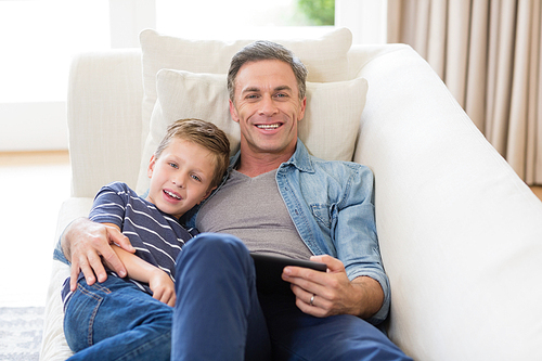 Portrait of father and son lying on sofa with digital tablet in living room at home