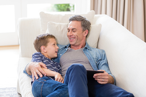 Smiling father and son lying on sofa with digital tablet in living room at home