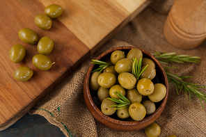 Overhead view of green olives with rosemary in bowl by cutting board on table