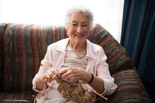 Portrait of smiling senior woman knitting while sitting on sofa against window at retirement home