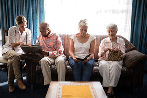 Smiling female doctor reading book by senior people sitting on sofa against window at retirement home