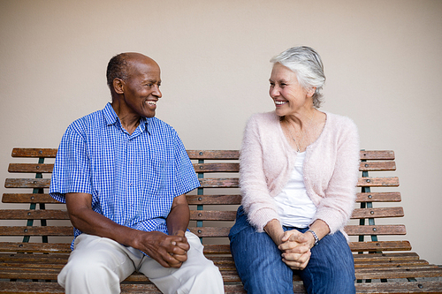 Smiling senior man and woman looking at each other while sitting on bench against wall at retirement home