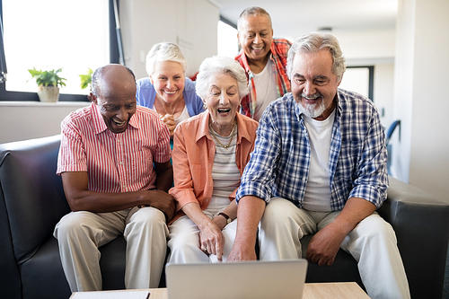 Cheerful senior friends looking at laptop on table while resting in nursing home