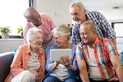 Senior woman showing digital tablet to cheerful friends while sitting on sofa at nursing home