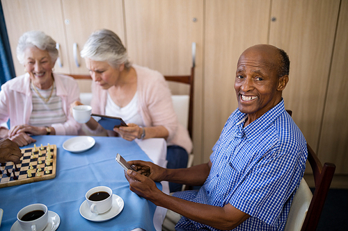 Portrait of senior man using mobile phone while having tea and playing chess with friends at table