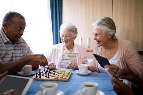 Happy senior friends playing chess while having coffee at table in nursing home