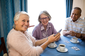 Smiling senior friends playing cards while having coffee at table in nursing home