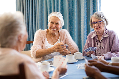 Smiling senior female friends playing cards while having coffee at table in nursing home
