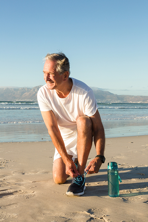 Smiling senior man tying shoelace at beach against clear sky