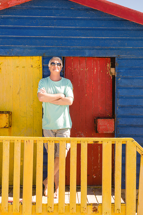 Senior man with arms crossed standing by railing of beach hut on sunny day