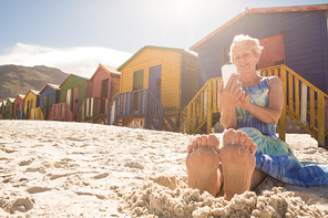 Senior woman using smart phone while sitting on sand against huts at beach