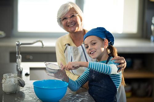 Grandmother posing with granddaughter sieving the flour in the kitchen