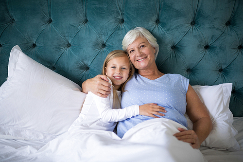 Portrait of smiling granddaughter and grandmother sitting together on bed in bedroom