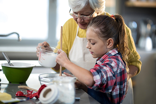 Grandmother adding water while granddaughter is mixing flour in a bowl in kitchen