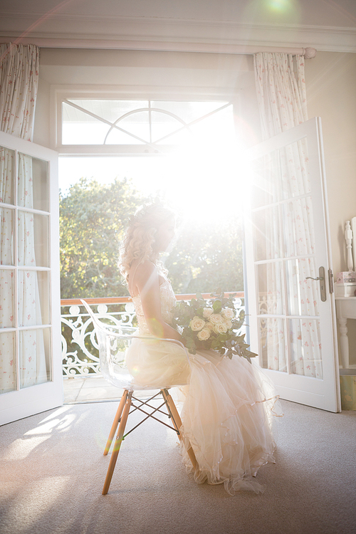 Side view of beautiful bride holding bouquet while sitting on chair at home