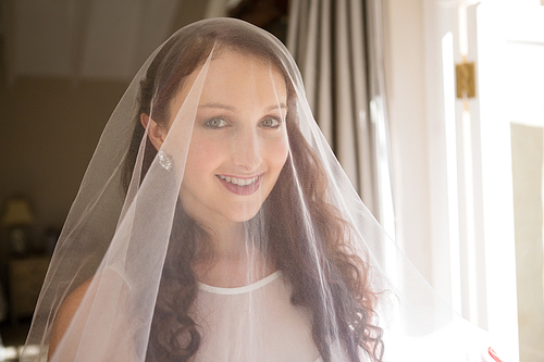 Portrait of happy bride in wedding dress standing by window at home