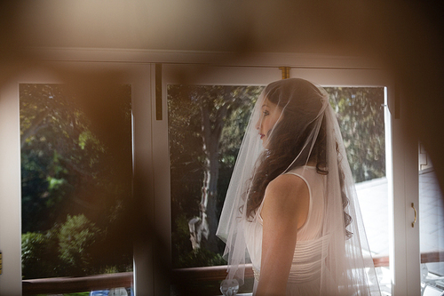 Beautiful bride in wedding dress looking through window at home