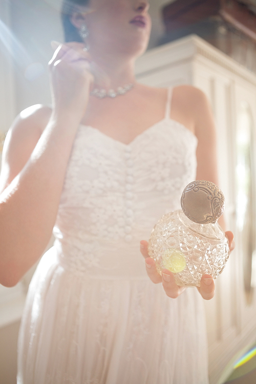 Low angle view of beautiful bride holding perfume while wearing earring at home