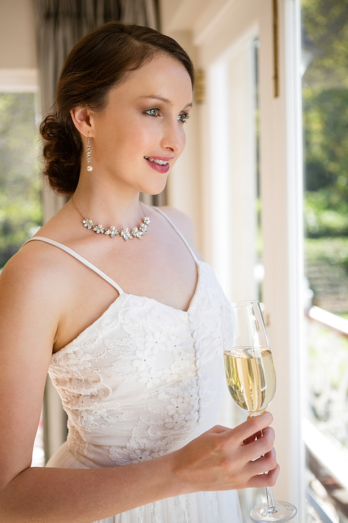 Bride holding champagne looking through window while standing at home