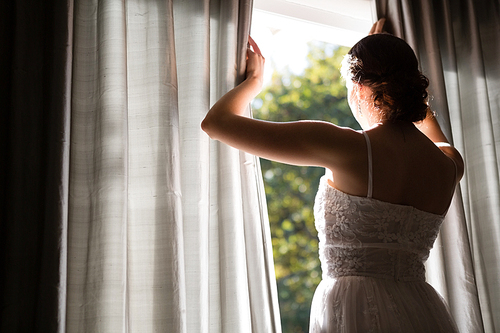 Rear view of bride in wedding dress looking through window while standing at home