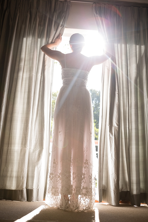 Low angle view of bride in wedding dress looking through window while standing at home