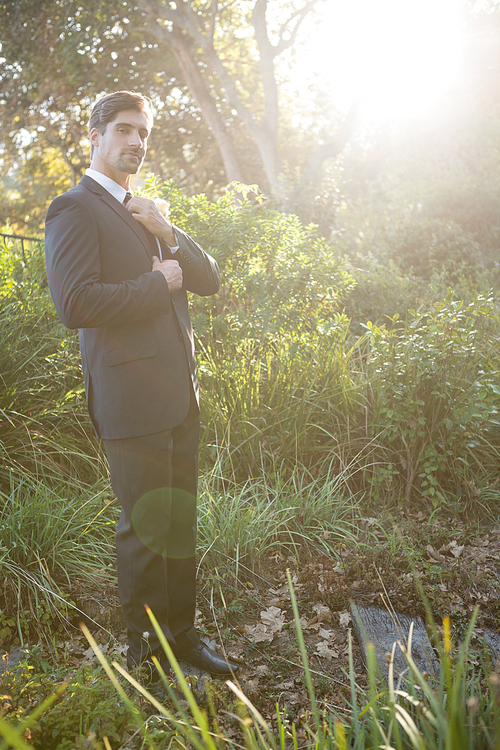 Portrait of confident bridegroom adjusting necktie while standing in park