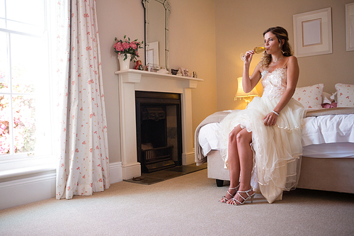 Bride drinking champagne while sitting on bed at home