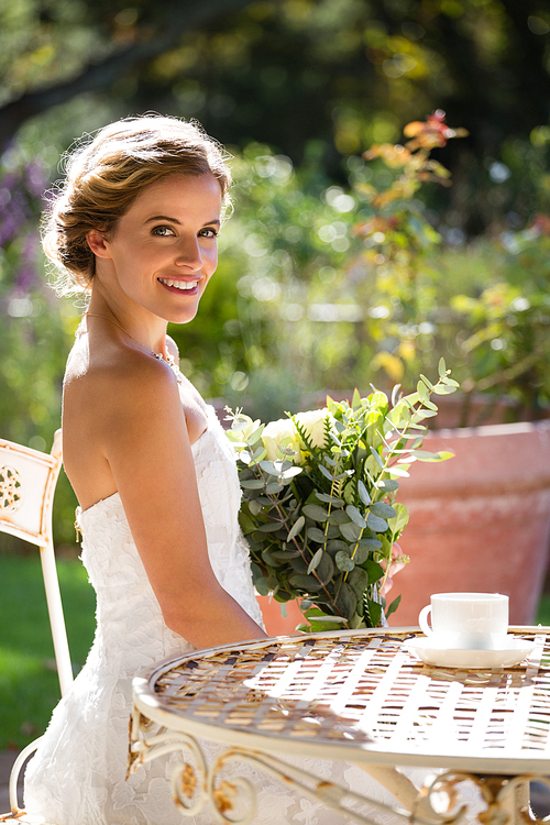 Portrait of smiling bride holding bouquet while sitting on chair in yard