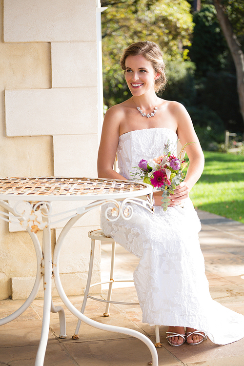 Beautiful bride with bouquet looking away while sitting on chair in yard