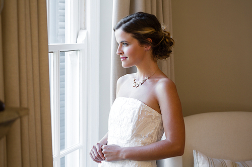 Thoughtful bride looking through window while standing at home
