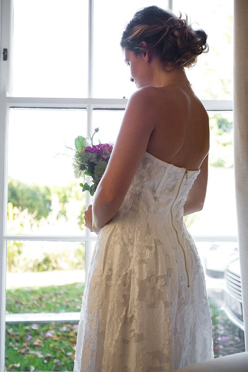 Side view of bride holding bouquet while looking through window at home