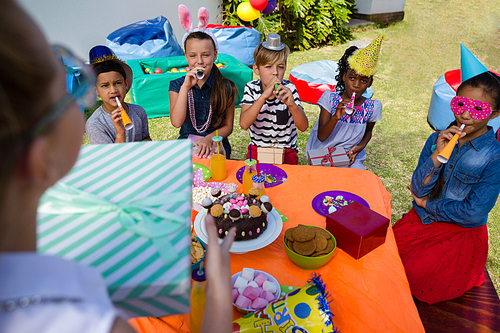 Girl holding gift while friends blowing party horn during birthday in yard