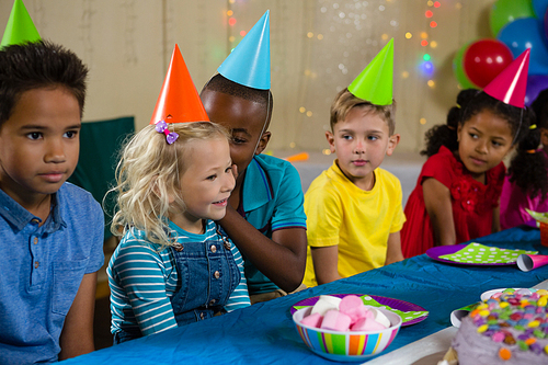 Friends looking at boy whispering to girl while sitting at table during birthday party
