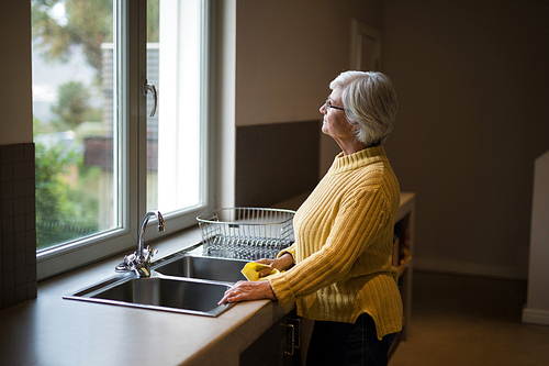 Thoughtful Senior woman standing near the kitchen sink and looking through window